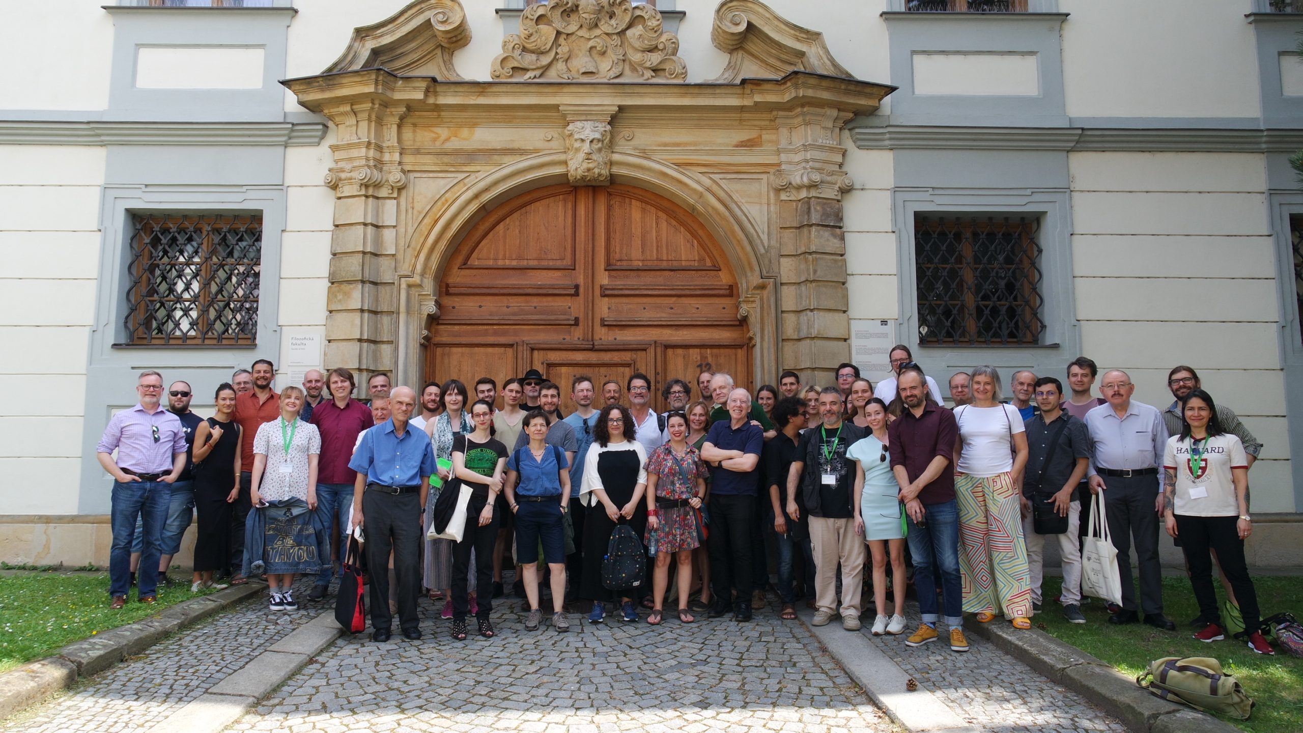 Group picture with around fifty people posing outdoors in front of a gigantic wooden door with a marble architrave of an old building.