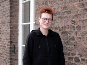 Portrait of a man wearing glasses and a black sweatshirt with zipper, smiling in front of a brick wall.