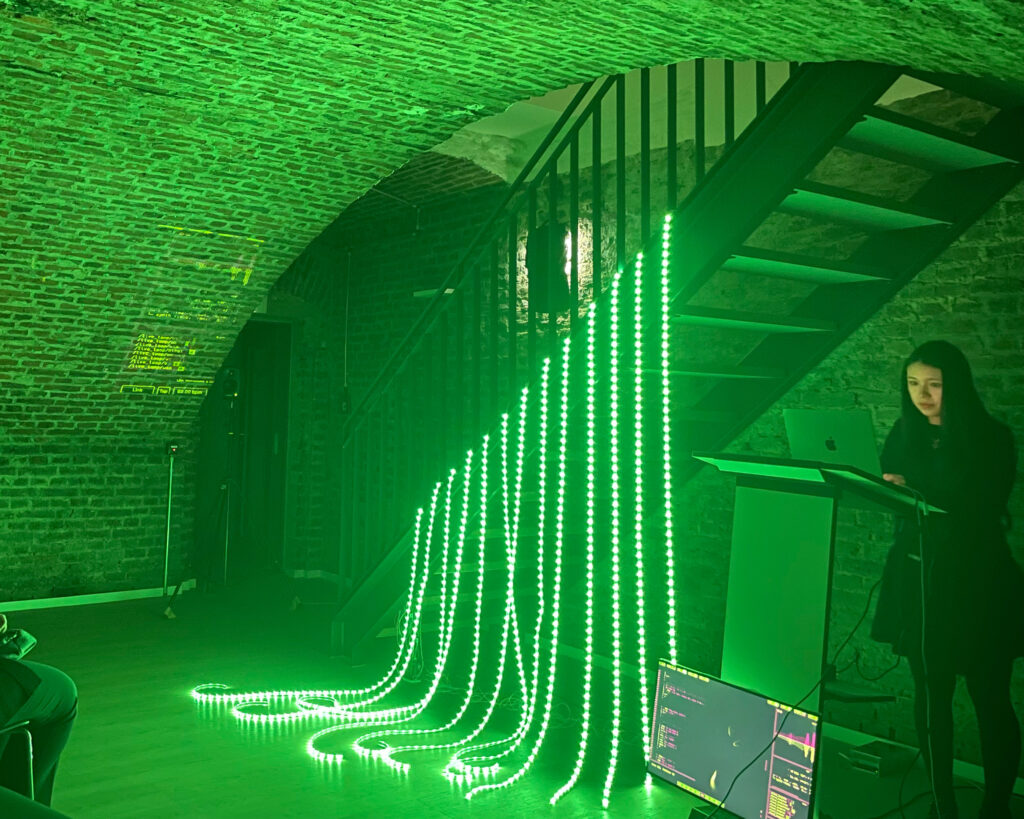 A female person standing behind a computer next to an art installation with green light tubes hanging on stairwell.