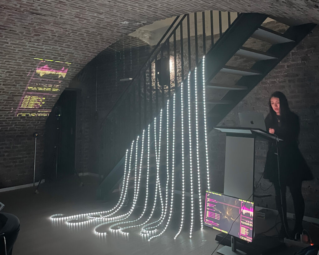 A female person standing behind a computer next to an art installation with white light tubes hanging on stairwell.