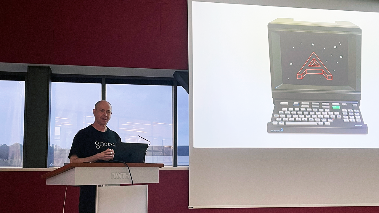 A man stands behind a speaker's desk next to a screen displaying an old computer.