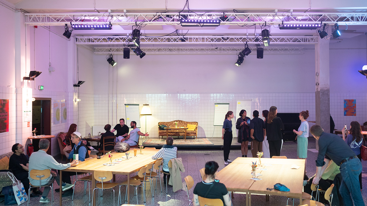 Sixteen people are standing or sitting in small groups, talking in a large room.