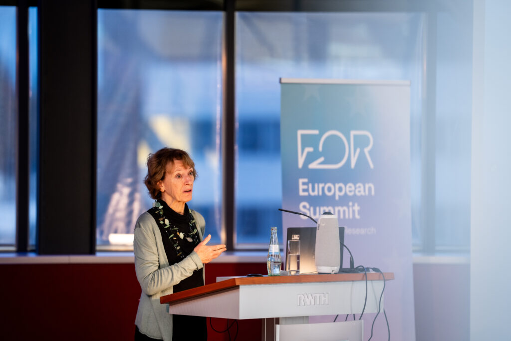 A woman standing on a desk. She has brown hair and wears a green cardigan with a black top and green, brown bluish necklace.