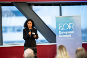 a woman with dark curly hair, wearing a black blazer and a skirt is talking, while holding a microphone.